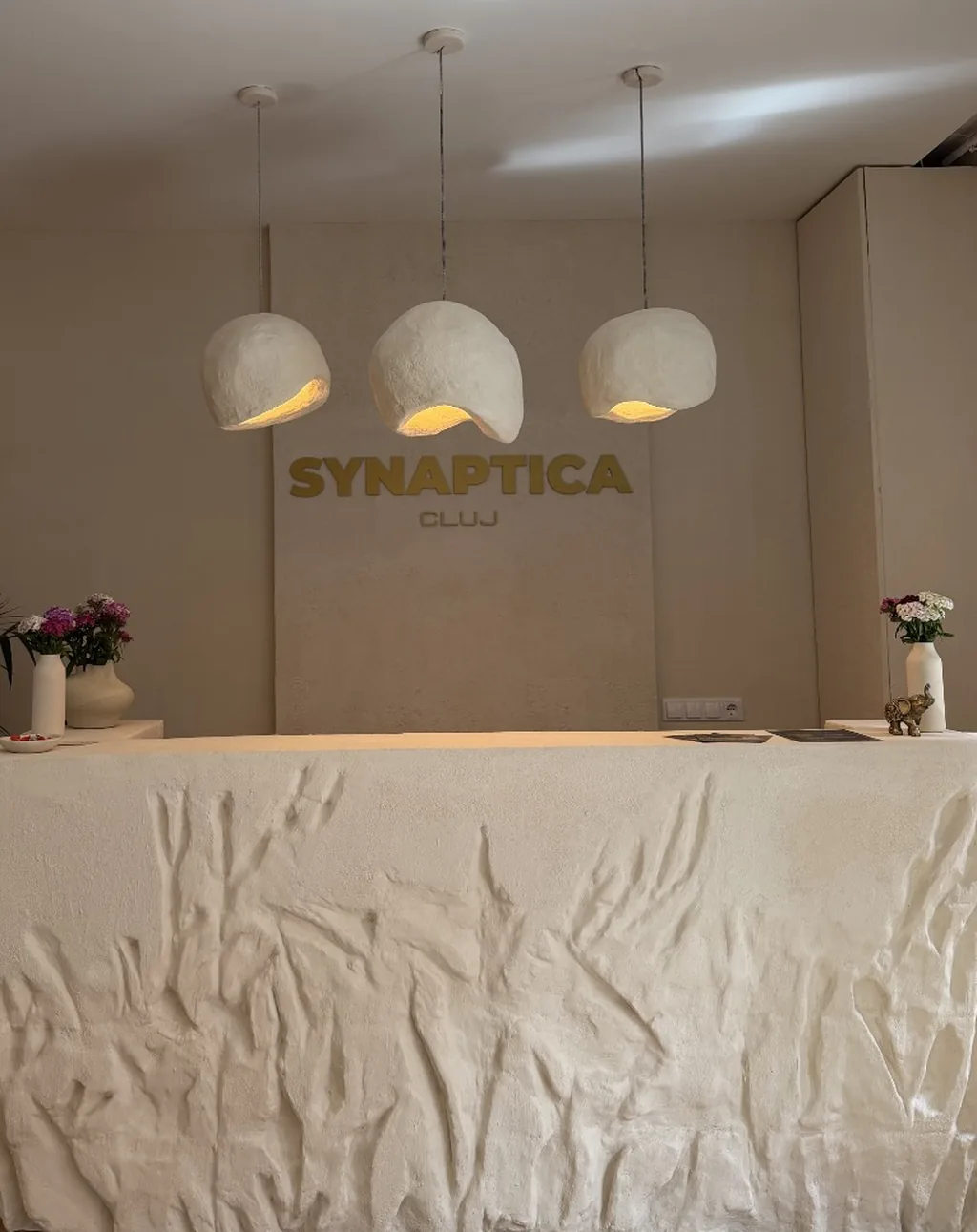Recepția Synaptica Cluj cu birou texturat alb, trei lămpi suspendate organice și branding auriu pe perete